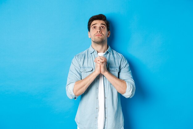 Jovem esperançoso olhando para cima e orando a Deus, implorando por ajuda, de pé contra um fundo azul.