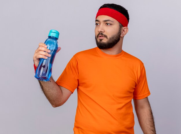 Jovem esportivo pensando, olhando para o lado usando bandana e pulseira segurando uma garrafa de água isolada no fundo branco