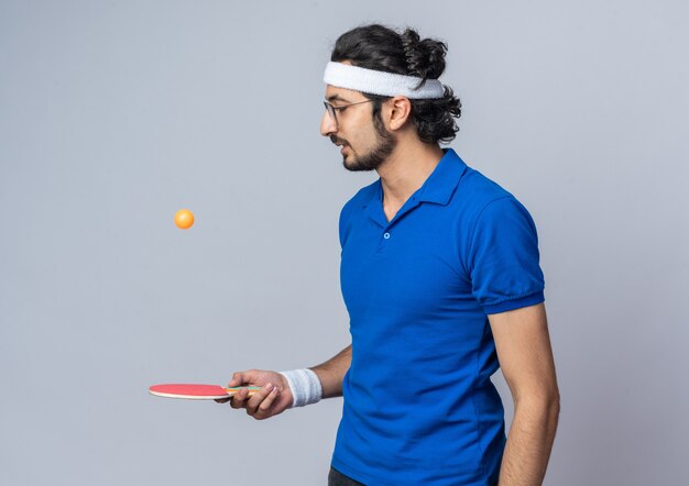 Jovem esportivo pensando usando bandana e pulseira segurando e olhando para a bola de pingue-pongue na raquete