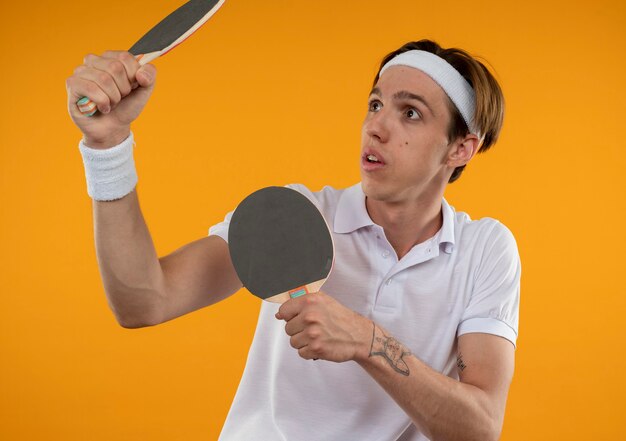 Jovem esportivo surpreso olhando para o lado usando bandana e pulseira levantando raquetes de pingue-pongue isoladas na parede laranja