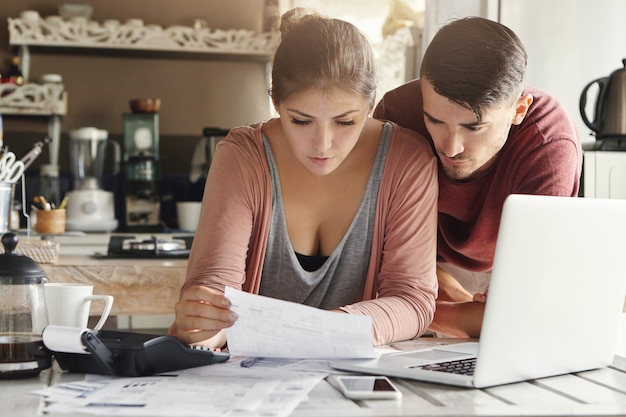 Foto grátis jovem esposa olhando um pedaço de papel com olhar sério, sentado à mesa da cozinha com laptop, calculadora e documentos