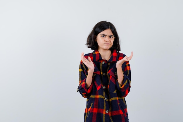 Jovem, esticando as mãos de forma questionadora em camisa quadriculada e olhando perplexa, vista frontal.