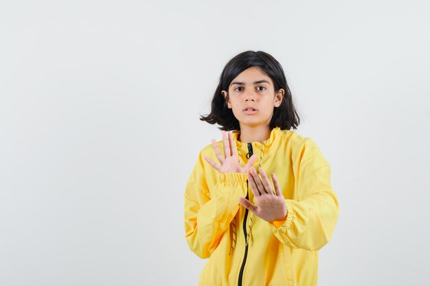 Jovem, esticando as mãos, mostrando um gesto de parada na jaqueta amarela e olhando séria