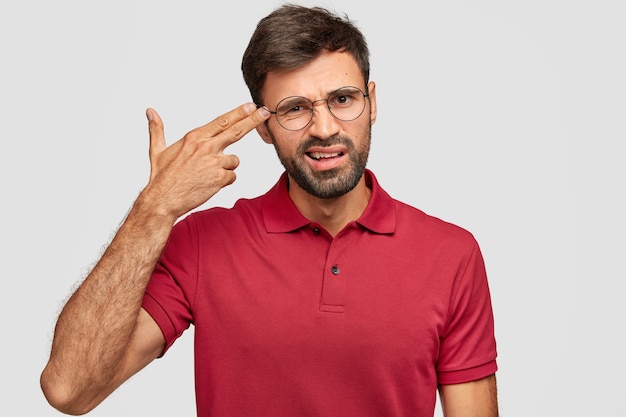 Jovem estressante faz gesto de arma, finge suicídio, mantém dois dedos nas têmporas, sente-se desesperado, posa sozinho contra uma parede branca, vestido com uma camiseta vermelha casual, tem crise