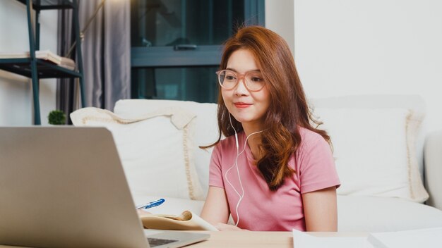 Jovem estudante adolescente da Ásia usa fones de ouvido, aula de aprendizagem à distância com o professor on-line no laptop na sala de estar de casa à noite. Distanciamento social, quarentena para prevenção do vírus corona.