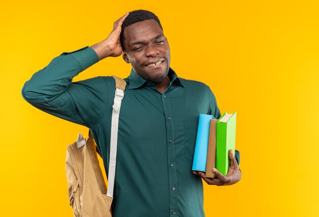 Jovem estudante afro-americana dolorida com uma mochila segurando livros e colocando a mão na cabeça, isolada em uma parede laranja com espaço de cópia