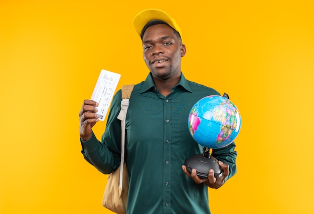 Jovem estudante afro-americana satisfeita com boné e mochila segurando a passagem aérea e o globo isolado na parede laranja com espaço de cópia