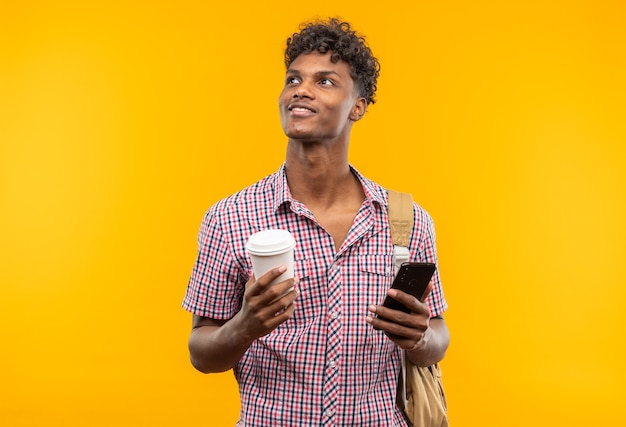 Jovem estudante afro-americana satisfeita com uma mochila segurando um telefone e um copo de papel olhando para o lado