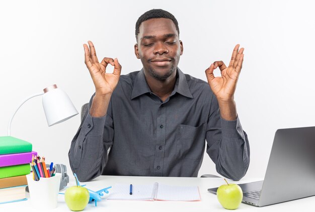 Jovem estudante afro-americana satisfeita, sentada com os olhos fechados na mesa com as ferramentas da escola, meditando isolada na parede branca