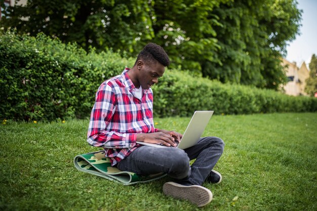Jovem estudante afro-americana sentada na grama com um laptop perto do campus
