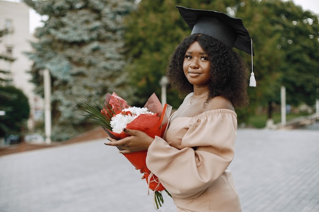 Jovem estudante afro americana vestida com um vestido bege e boné acadêmico. Campus como pano de fundo