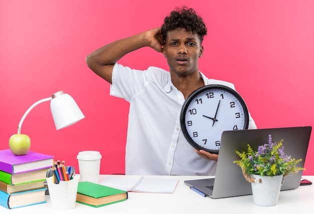 Jovem estudante afro-americano ansioso sentado na mesa com as ferramentas da escola, colocando a mão na cabeça dele e segurando o relógio