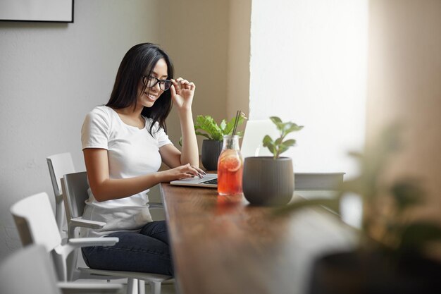 Jovem estudante asiático usando laptop no café ou coworking bebendo limonada sentado ao lado de uma grande janela Conceito de estudo