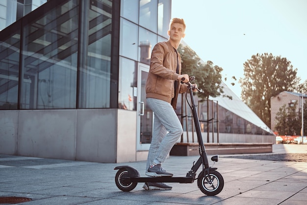 Jovem estudante atraente está dirigindo seu edifício de vidro interessante de scooter em dia ensolarado.