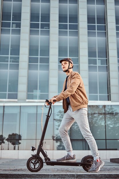 Jovem estudante atraente está dirigindo seu edifício de vidro interessante de scooter em dia ensolarado.