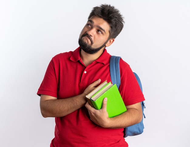 Jovem estudante barbudo com uma camisa pólo vermelha com mochila segurando cadernos olhando para cima com expressão cética no rosto em pé sobre um fundo branco