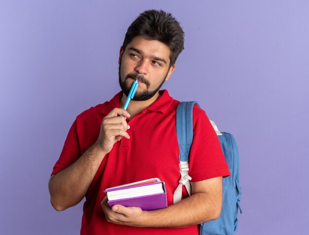 Jovem estudante barbudo com uma camisa pólo vermelha com uma mochila segurando livros e uma caneta olhando de lado perplexo em pé