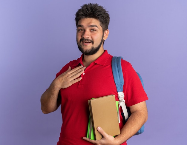 Jovem estudante barbudo com uma camisa pólo vermelha com uma mochila segurando livros, segurando a mão no peito, sentindo-se grato sorrindo alegremente em pé sobre a parede azul