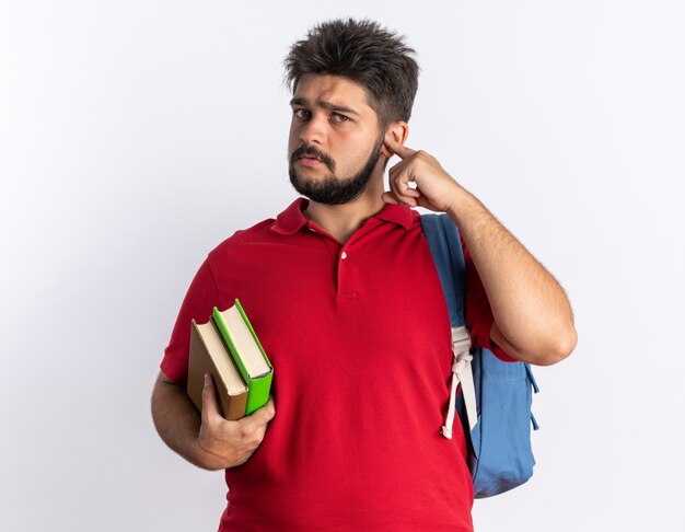 Jovem estudante barbudo em uma camisa pólo vermelha com mochila segurando cadernos parecendo intrigado com uma cara séria em pé