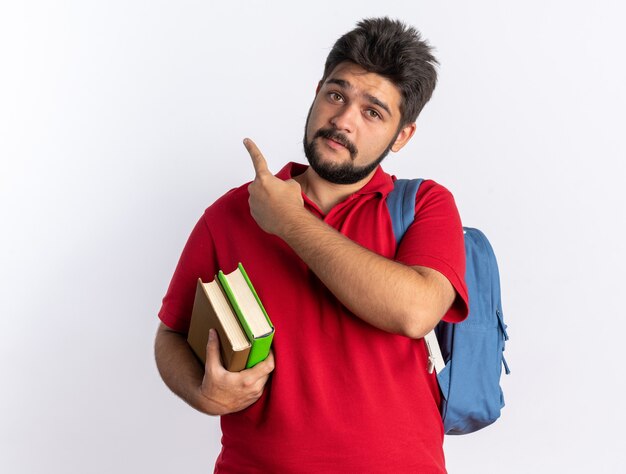 Jovem estudante barbudo em uma camisa pólo vermelha com mochila segurando notebooks lookign para a câmera feliz e positivo apontando com o dedo indicador para o lado em pé sobre um fundo branco