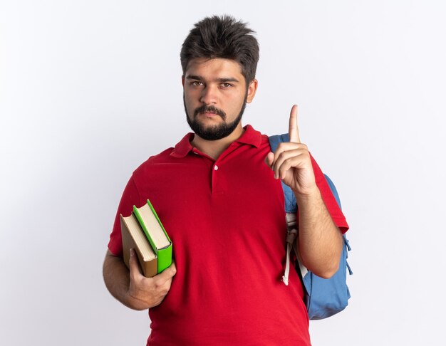 Jovem estudante barbudo em uma camisa pólo vermelha com mochila segurando notebooks olhando para a câmera com uma cara séria mostrando o dedo indicador em pé sobre um fundo branco