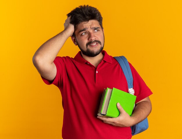 Foto grátis jovem estudante barbudo em uma camisa pólo vermelha com mochila segurando notebooks parecendo confuso com a mão na cabeça por engano em pé