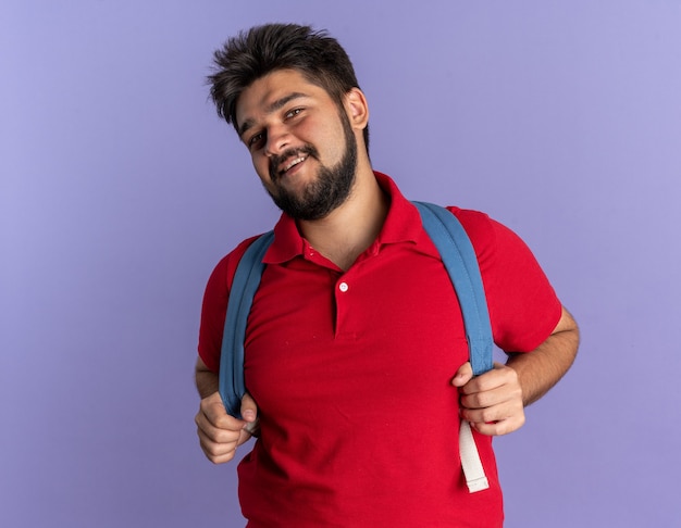 Jovem estudante barbudo em uma camisa pólo vermelha com mochila, sorrindo, confiante, feliz e positivo