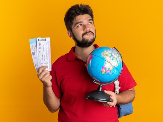 Jovem estudante barbudo em uma camisa pólo vermelha com uma mochila segurando um globo e as passagens aéreas, olhando para cima intrigado e feliz em pé