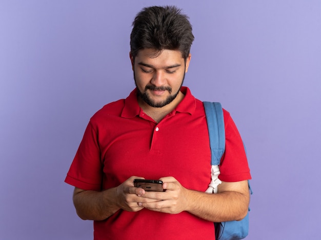 Jovem estudante barbudo em uma camisa pólo vermelha com uma mochila segurando um smartphone escrevendo uma mensagem de texto, parecendo confiante em pé