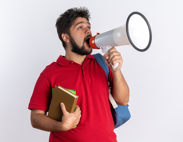 Jovem estudante barbudo emocional em uma camisa pólo vermelha com mochila segurando notebooks gritando para o megafone em pé sobre a parede branca
