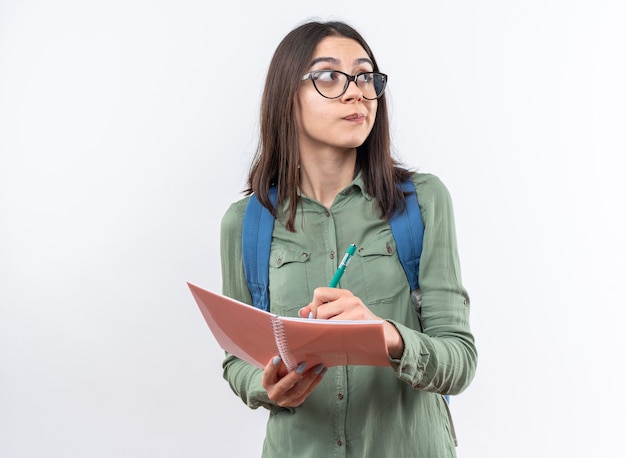 Jovem estudante de óculos com mochila a pensar e a escrever algo no caderno