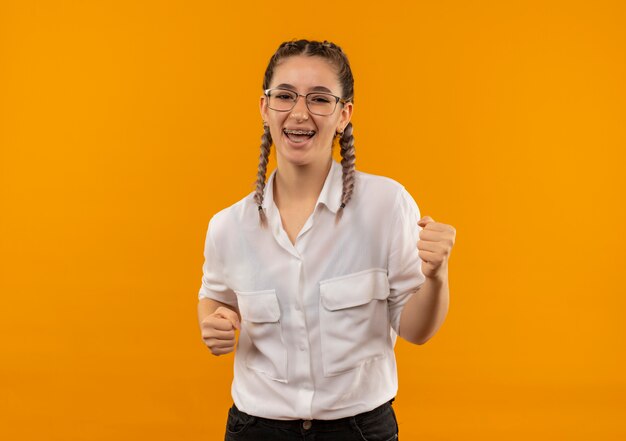 Jovem estudante de óculos com rabo de cavalo na camisa branca cerrando os punhos feliz e animada, regozijando-se com seu sucesso em pé sobre a parede laranja