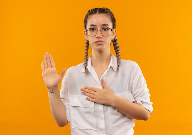 Jovem estudante de óculos com rabo de cavalo na camisa branca olhando para a frente fazendo um juramento ou fazendo promessa em pé sobre uma parede laranja