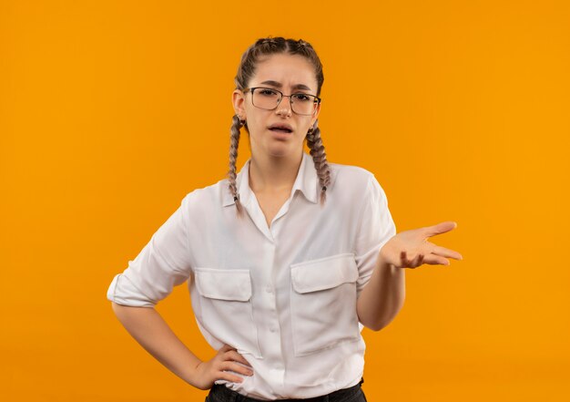Jovem estudante de óculos com rabo de cavalo na camisa branca parecendo descontente com o braço estendido enquanto faz uma pergunta em pé sobre a parede laranja
