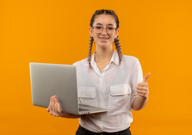 Jovem estudante de óculos com rabo de cavalo na camisa branca segurando um laptop olhando para a frente sorrindo e mostrando os polegares em pé sobre a parede laranja