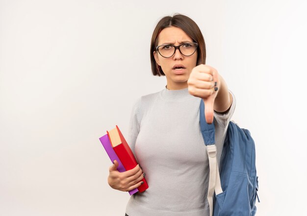 Jovem estudante descontente usando óculos e uma bolsa com as costas segurando livros, mostrando o polegar para baixo, isolado no fundo branco com espaço de cópia