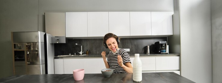 Foto grátis jovem estudante despreocupada com fones de ouvido a comer cereais ao pequeno-almoço a ouvir música a cantar e