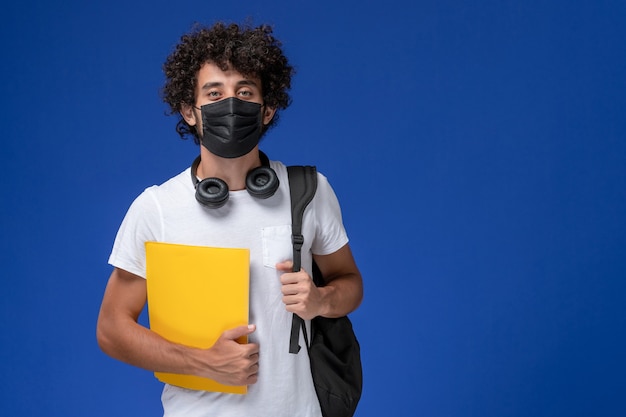 Jovem estudante do sexo masculino de vista frontal em t-shirt branca usando máscara preta e segurando arquivos amarelos sobre o fundo azul claro.