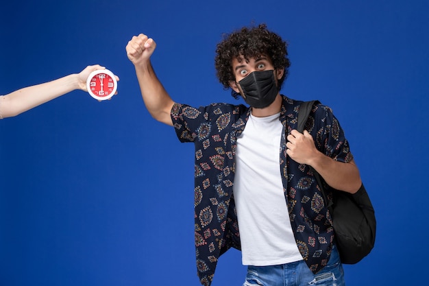 Jovem estudante do sexo masculino de vista frontal usando máscara preta com mochila em fundo azul claro.