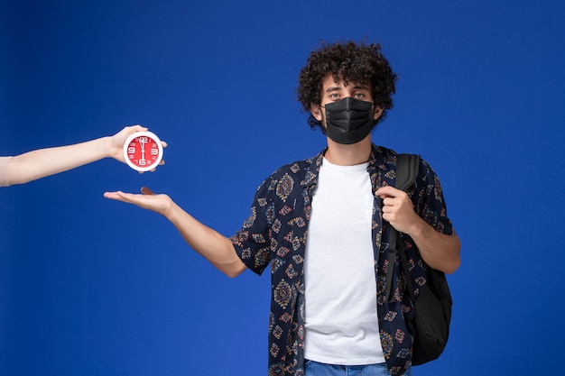 Jovem estudante do sexo masculino de vista frontal usando máscara preta com mochila em fundo azul claro.