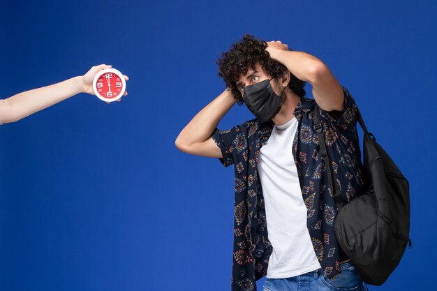 Jovem estudante do sexo masculino de vista frontal usando máscara preta com mochila posando em fundo azul claro.