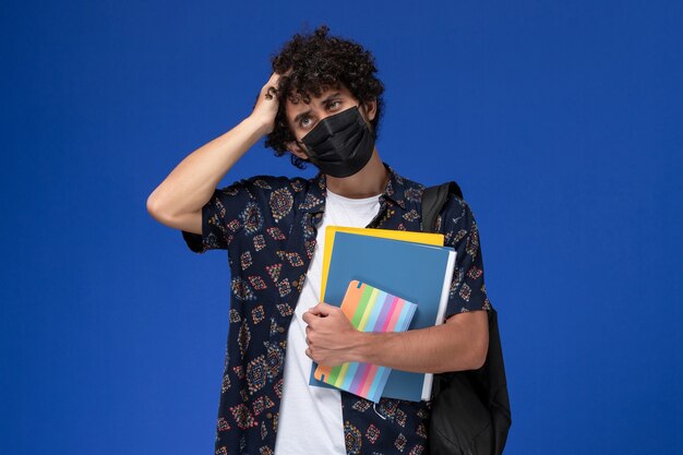 Jovem estudante do sexo masculino de vista frontal usando máscara preta com mochila segurando arquivos e caderno pensando sobre o fundo azul.