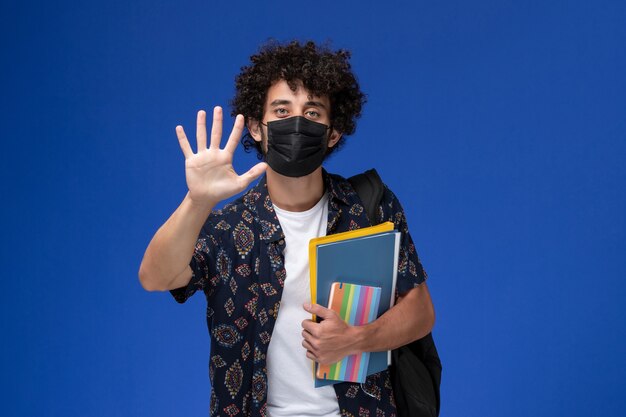 Jovem estudante do sexo masculino de vista frontal usando máscara preta com mochila segurando o caderno e arquivos na mesa azul.