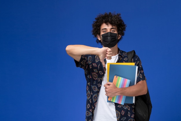 Jovem estudante do sexo masculino de vista frontal usando máscara preta com mochila segurando o caderno e arquivos sobre fundo azul.