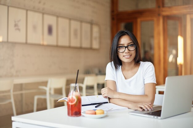 Jovem estudante empresário asiático trabalhando no laptop na biblioteca ou café de espaço aberto, olhando para a câmera sorrindo.