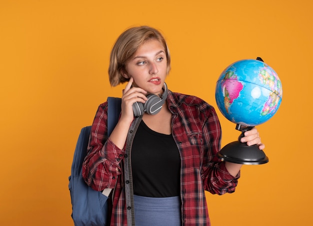 Foto grátis jovem estudante eslava confusa com fones de ouvido e mochila colocando o dedo no rosto segurando e olhando para o globo