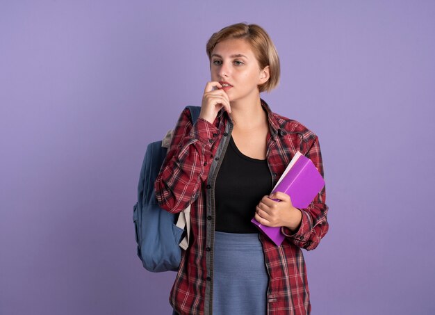 Jovem estudante eslava confusa usando mochila morde unha segura livro e caderno