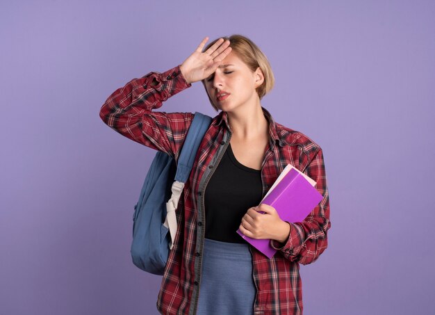 Jovem estudante eslava dolorida usando uma mochila e colocando a mão na testa segurando um livro e um caderno