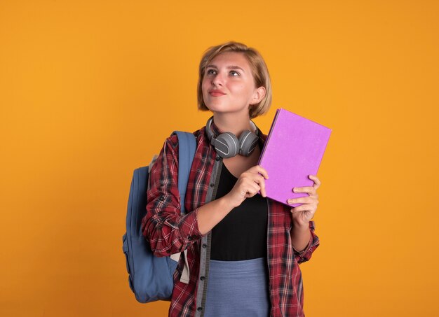 Jovem estudante eslava impressionada com fones de ouvido usando uma mochila segurando um livro e um caderno olhando para o lado