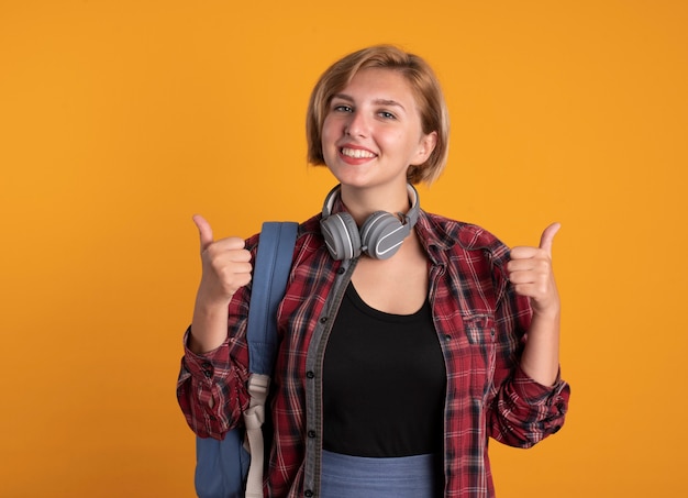 Foto grátis jovem estudante eslava sorridente com fones de ouvido e mochila com os polegares para cima com as duas mãos